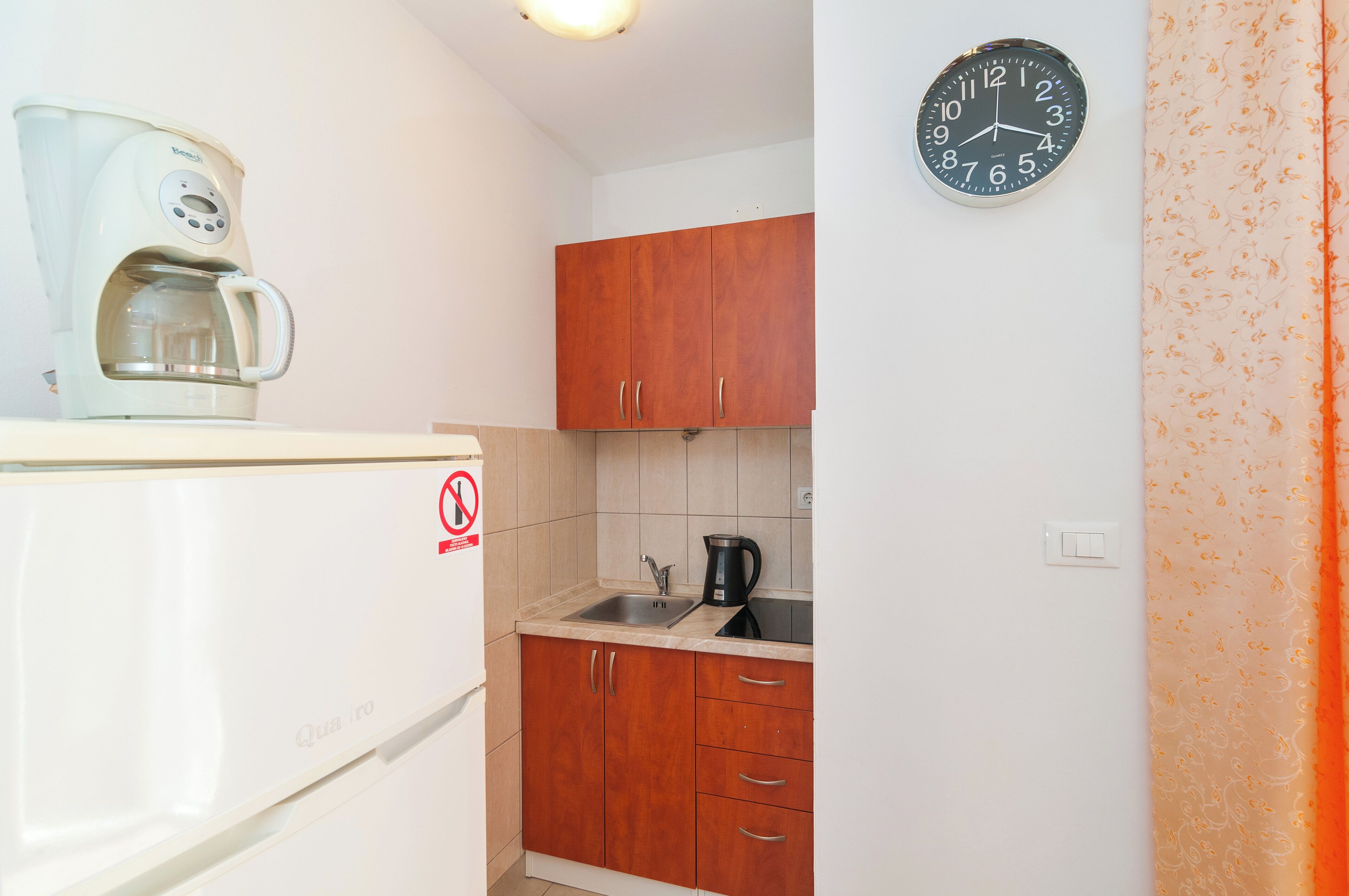 Una cucina compatta con una macchina per il caffè, un bollitore nero, mobili in legno e una sveglia a parete.