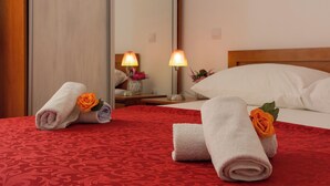 Una habitación de hotel con una colcha estampada roja, dos toallas enrolladas con rosas naranjas, un cabecero de madera, una mesita de noche con una lámpara y un espejo que refleja la habitación.