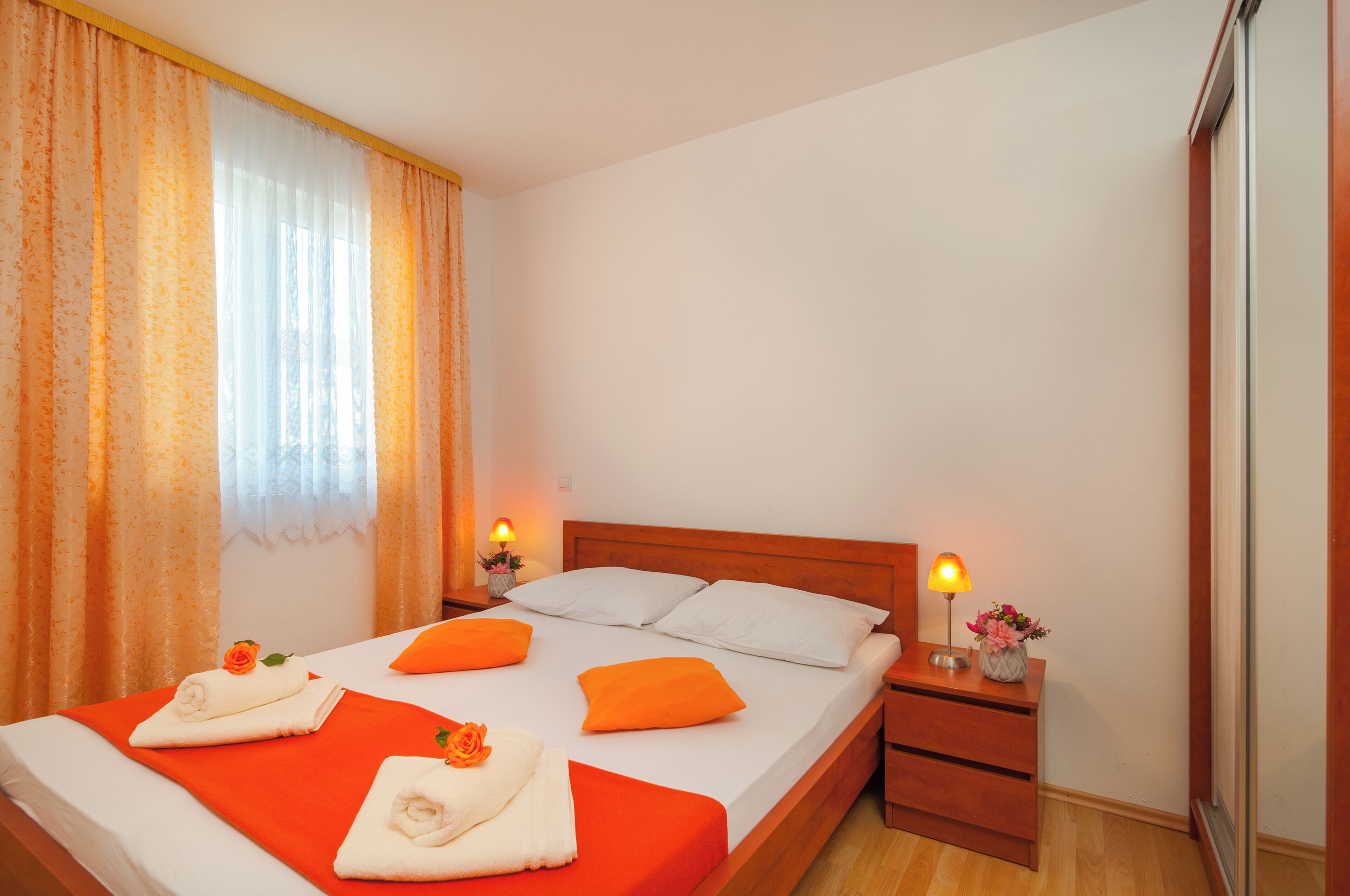 A neatly made bed with orange bedding, white pillows, and a wooden headboard. There are two bedside tables with lamps and flower arrangements.