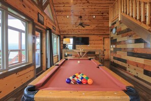 A pool table with balls in a wooden room with large windows and a television.