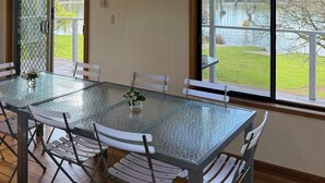 A dining area with a glass table and white chairs, a hanging light fixture, and a view of a lake through large windows. - Golden Mile - On the Waterfront (Bundalong)