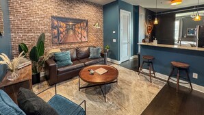 Un salon moderne avec un mur en briques, un canapé en cuir, une table basse ronde et un coin cuisine avec des tabourets.