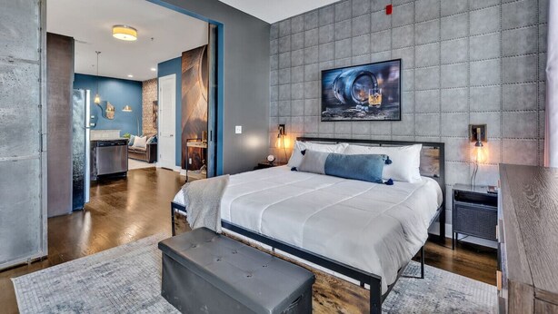 Une chambre à coucher moderne avec un grand lit, une tête de lit agrémentée d’un mur texturé et un tableau au-dessus. On y trouve une petite table de chevet avec une lampe et une prise de courant.