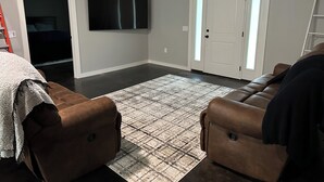 Un salon doté d’un grand téléviseur à écran plat fixé au mur, d’un canapé en cuir marron, d’un tapis à motifs et d’une porte avec une fenêtre.