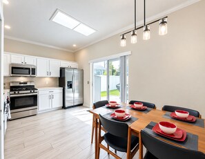 Cocina moderna con electrodomésticos de acero inoxidable, zona de comedor con mesa de madera y sillas negras, y puerta corrediza de vidrio que da a un patio trasero.