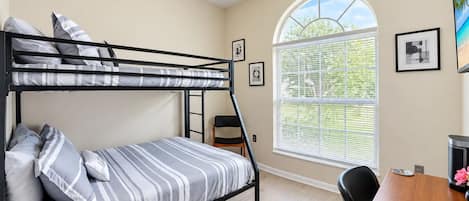 A bunk bed with striped bedding, a desk with a chair, a window with blinds, and framed pictures on the wall.