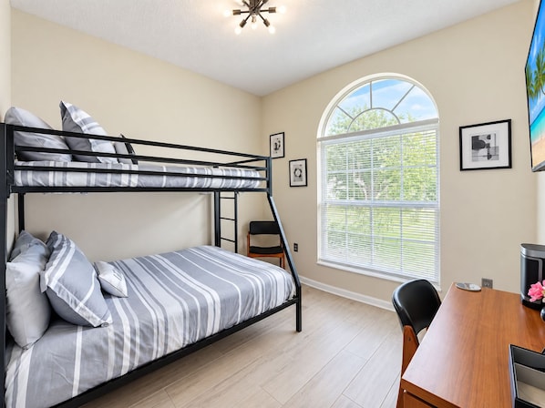 A bunk bed with striped bedding, a desk with a chair, a window with blinds, and framed pictures on the wall.