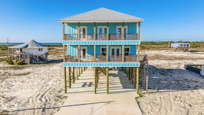 Une maison de plage à deux étages, dotée d’un balcon, d’un chemin en béton menant à l’entrée et, en arrière-plan, d’une plage de sable.