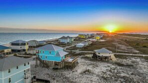 Zona residencial costera con casas sobre pilotes, una playa y un atardecer.