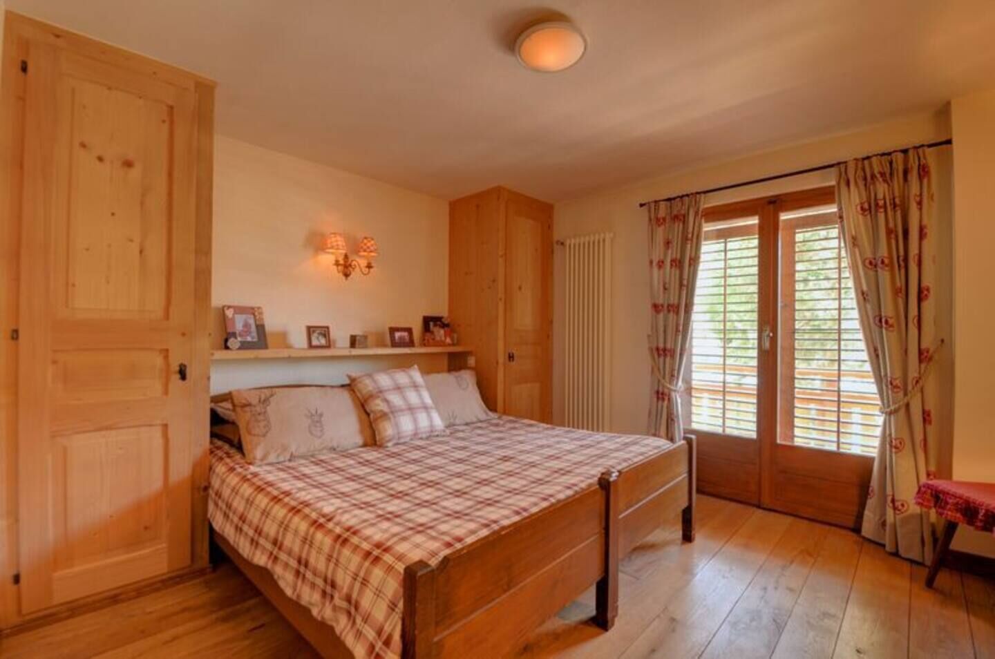 A bedroom with a wooden bed, a nightstand, a shelf with framed photos, and a window with curtains.