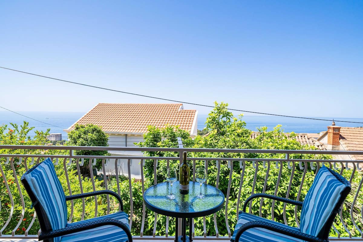 A balcony with two chairs and a table, overlooking a coastal area with houses and a clear sky.