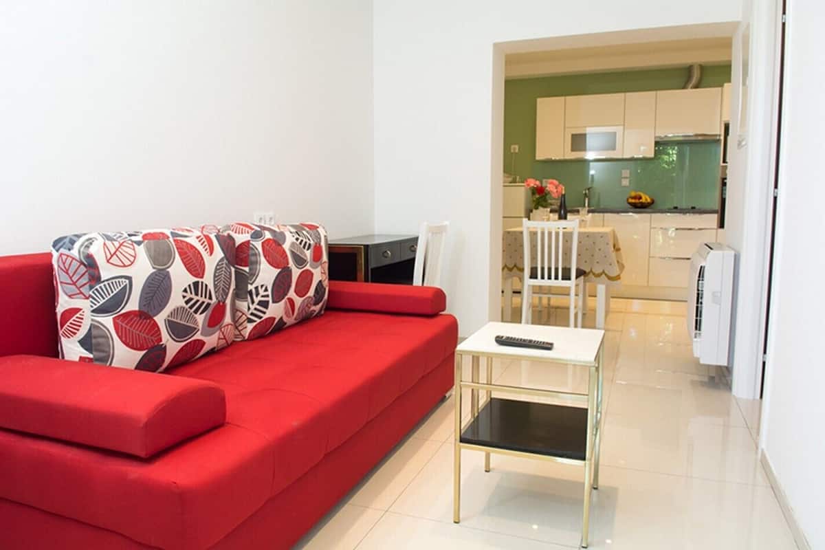 A red sofa with patterned pillows, a small white table with a remote control, and a kitchen area with white cabinets and a microwave.