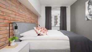 Un dormitorio moderno con pared de ladrillo, una cama con ropa de cama blanca y naranja, una mesita de noche con lámpara y vista a la calle a través de una ventana.