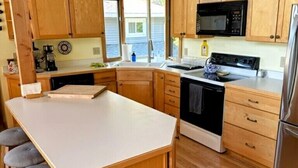 Cocina con armarios de madera, encimera blanca, microondas, horno y refrigerador. Hay taburetes en la isla de la cocina.