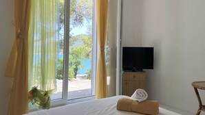A hotel room with a bed, a TV on a wooden stand, and a view of the sea through a large window. - Apartments More & Ja (Ston)