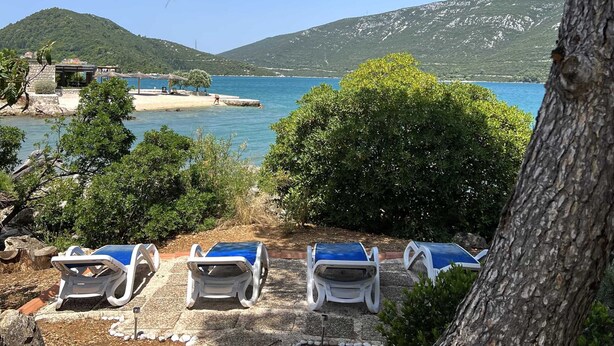 A coastal view with a clear sky, a beach, and a mountain in the background. There are lounge chairs and a small building near the water. - Apartments More & Ja (Ston)
