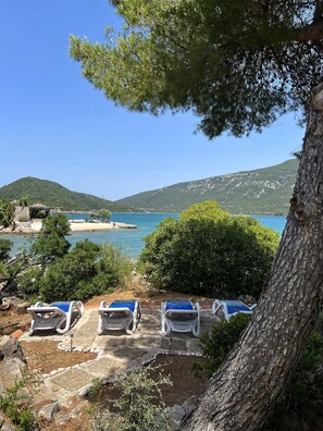 A coastal view with a clear sky, a beach, and a mountain in the background. There are lounge chairs and a small building near the water. - Apartments More & Ja (Ston)