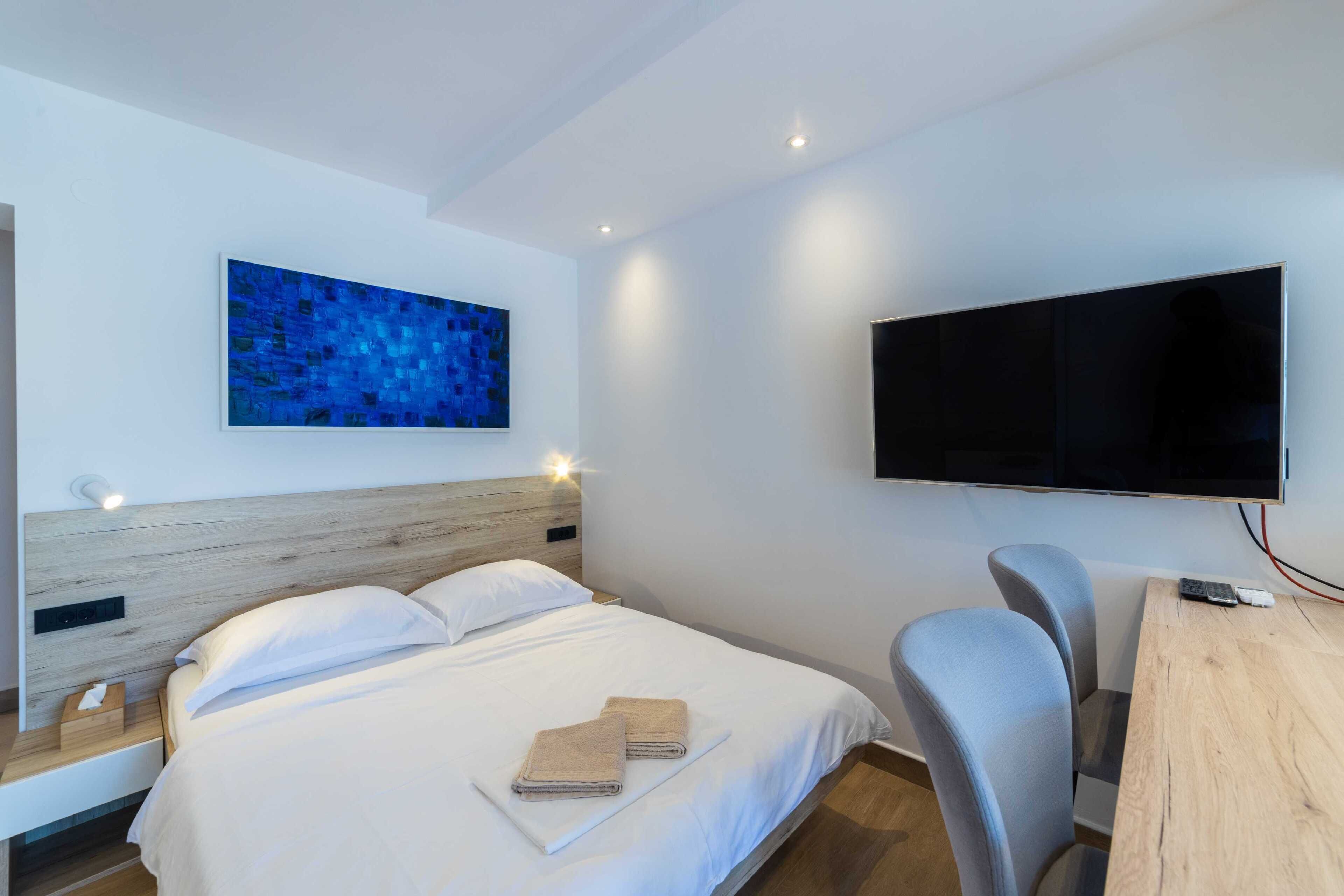 Ein modernes Hotelzimmer mit einem großen Bett, einem Flachbildfernseher, einem Schreibtisch mit Stuhl und einem blauen, abstrakten Gemälde an der Wand.