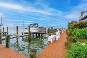 A wooden dock leading to a marina with boats, a building with a balcony, and a well-maintained garden.
