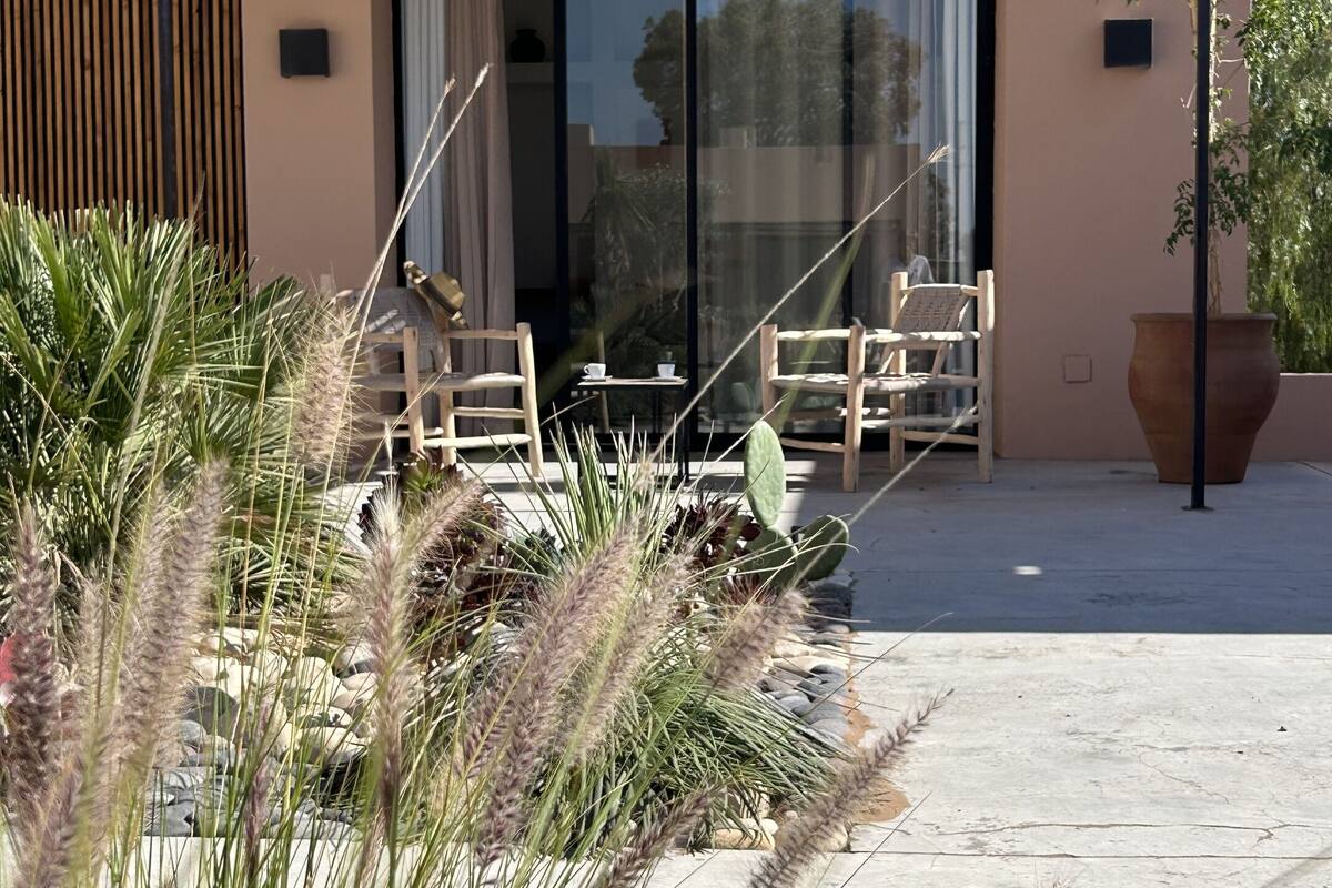 A modern building with a glass door entrance, outdoor seating area with chairs and tables, and a thatched roof structure.