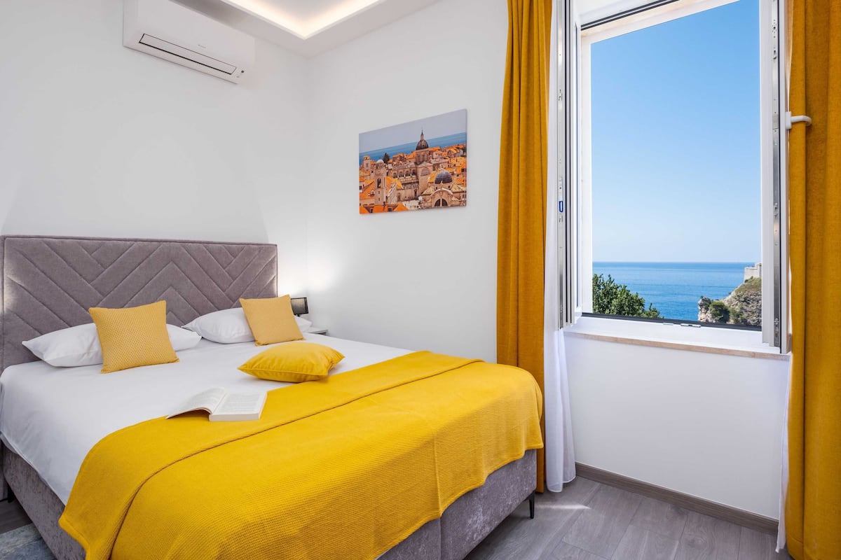 Een slaapkamer met een bed, een nachtkastje, een wandairconditioner, een raam met uitzicht op zee en een schilderij van een kustplaats.