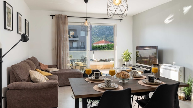 A modern living room with a sofa, dining table, and a TV. The room has a large window with a view of a building and greenery. - Apartment Why Not (Dubrovnik)