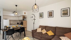 A modern living room with a brown sofa, a glass coffee table, and a dining area with a wooden table and chairs. There are framed pictures on the wall and a pendant light above the dining table. - Apartment Why Not (Dubrovnik)