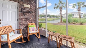 Une véranda avec un mur en briques, une porte blanche, deux fauteuils à bascule, une petite table avec une plante, et une vue sur un lac et des palmiers.