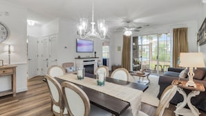 Un coin repas avec une table en bois, des chaises et un lustre. Un téléviseur est fixé au mur et un ventilateur de plafond surplombe l’espace détente.