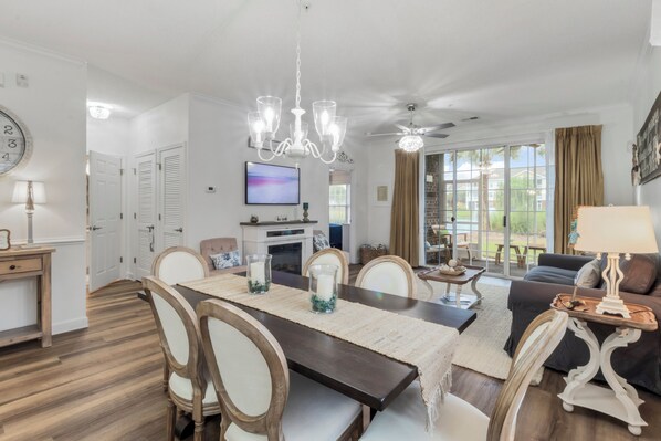 A dining area with a wooden table, chairs, and a chandelier. There is a television mounted on the wall and a ceiling fan above the seating area.
