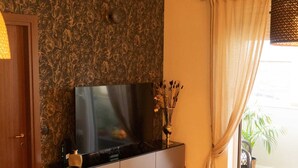 Una sala de estar con un televisor de pantalla plana sobre un mueble oscuro, un sillón verde, una mesita redonda con velas y una ventana con cortinas.