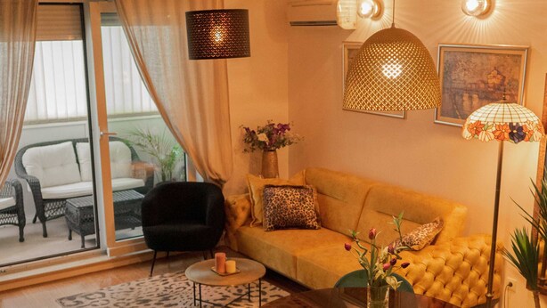 Una sala de estar moderna con un sofá, sillas verdes, una mesa de centro y una mesa de comedor. Hay una ventana grande con cortinas, una lámpara de pie y un aplique de pared.