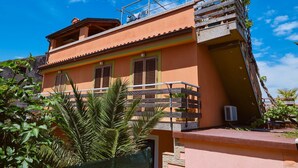 A two-story building with a balcony, a small garden area, and a clear blue sky. - Apartment Blamax (Mali Losinj)