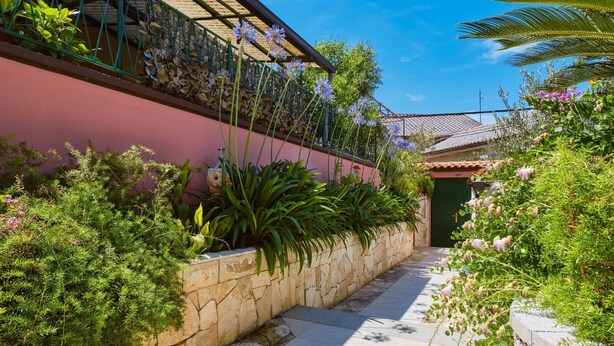 A narrow stone pathway flanked by lush greenery and a pink building with a tiled roof. - Apartment Blamax (Mali Losinj)