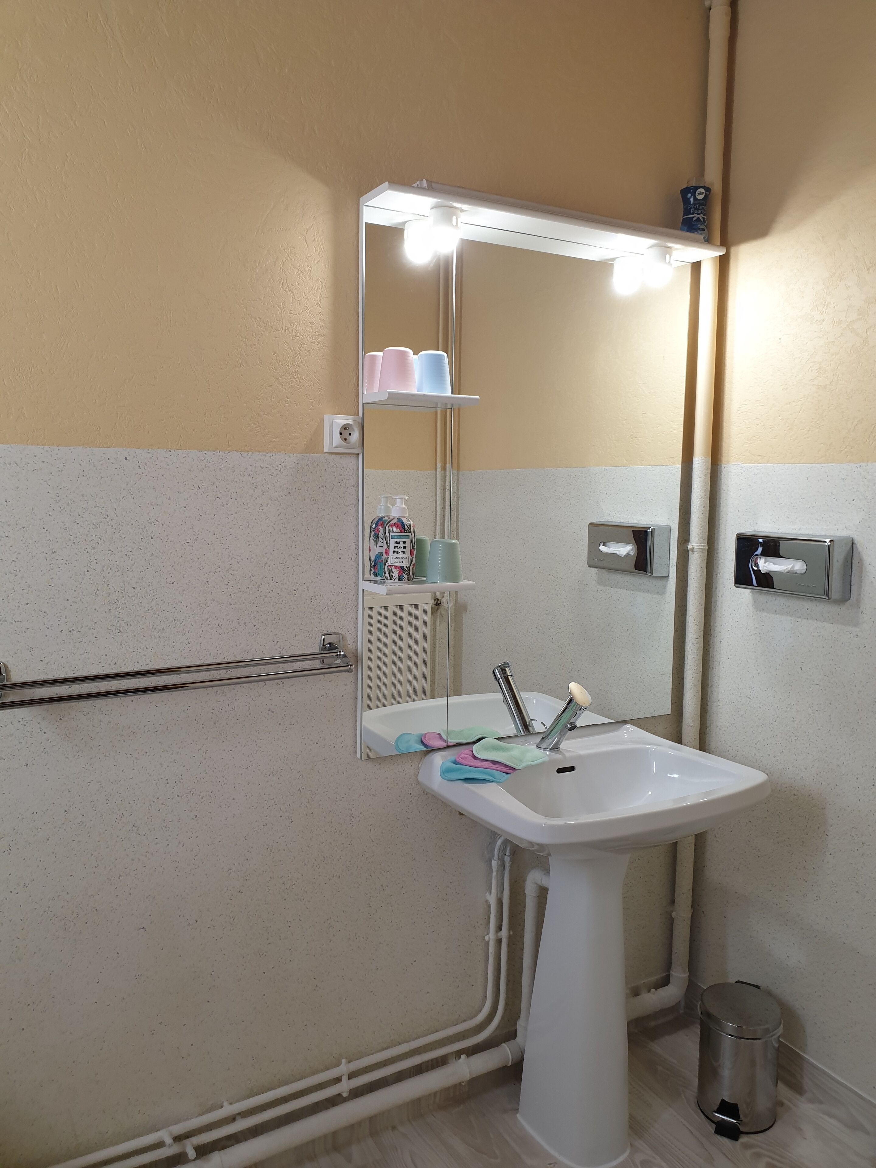A bathroom with a white sink, a mirror with lights, a towel rack, and a trash can.
