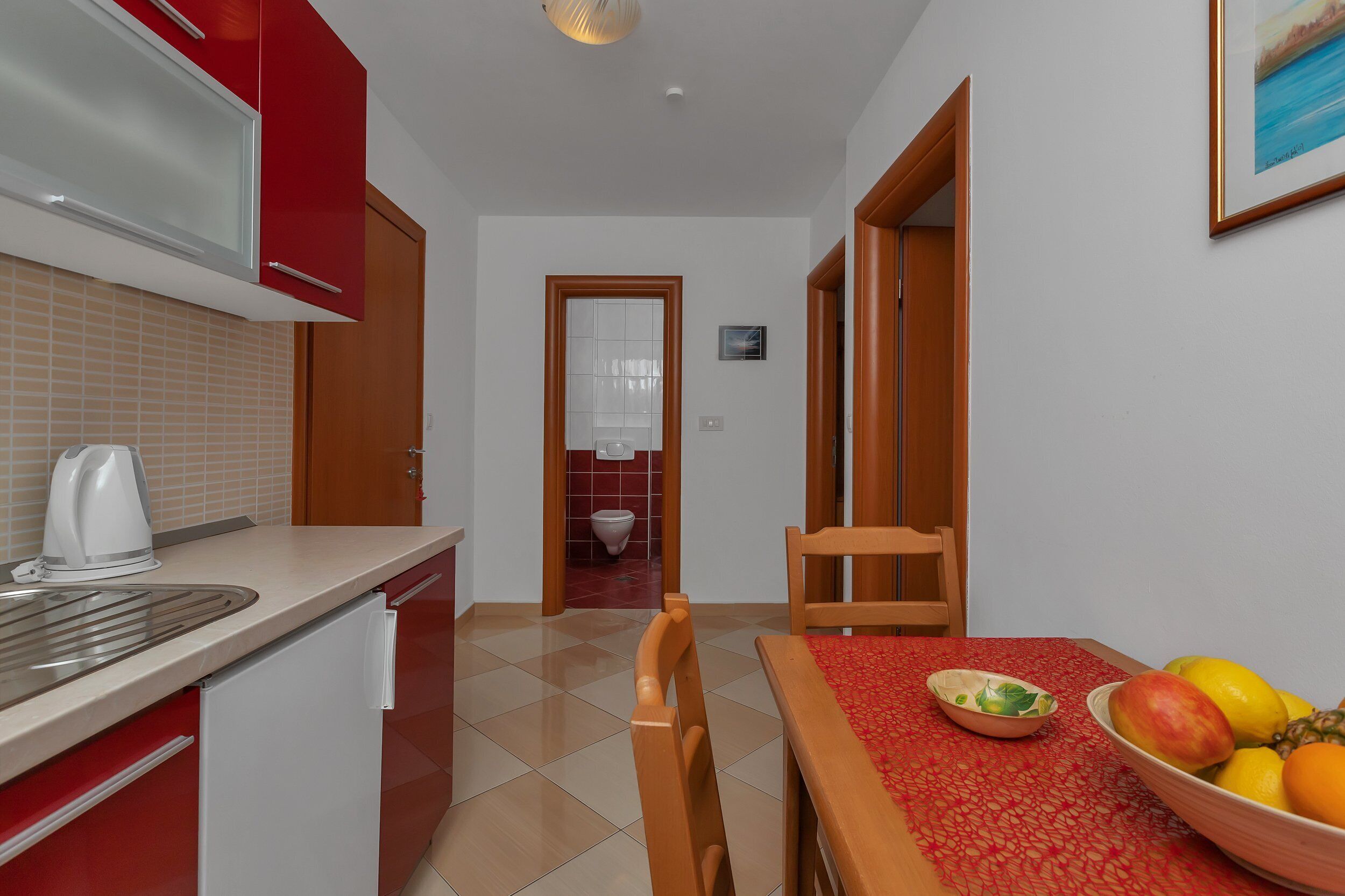 A compact kitchen with a red and white color scheme, a sink, a kettle, and a bowl of fruit on the table.