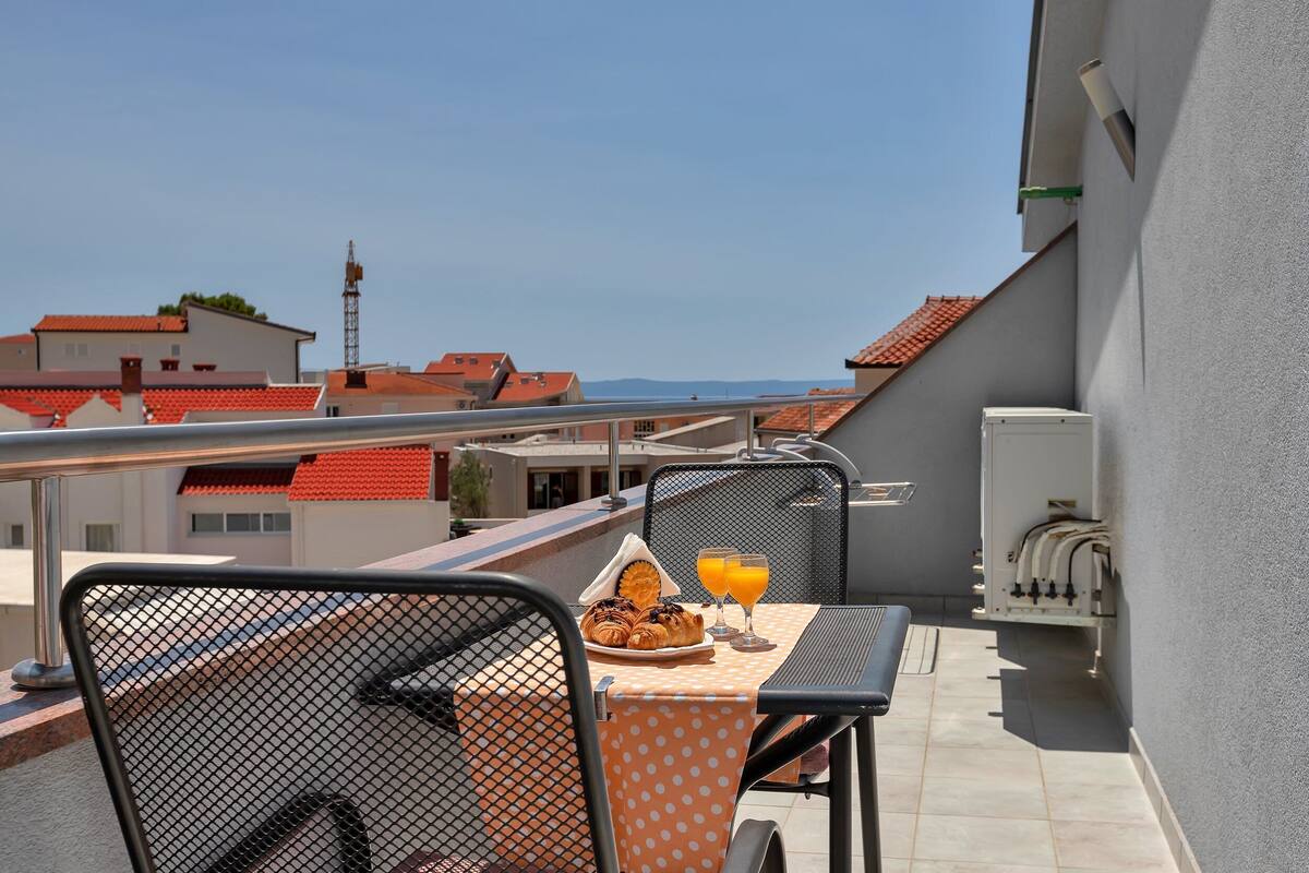 Een balkon met een ontbijttafel, compleet met croissants en sinaasappelsap, met uitzicht op een stadsbeeld met rode daken.