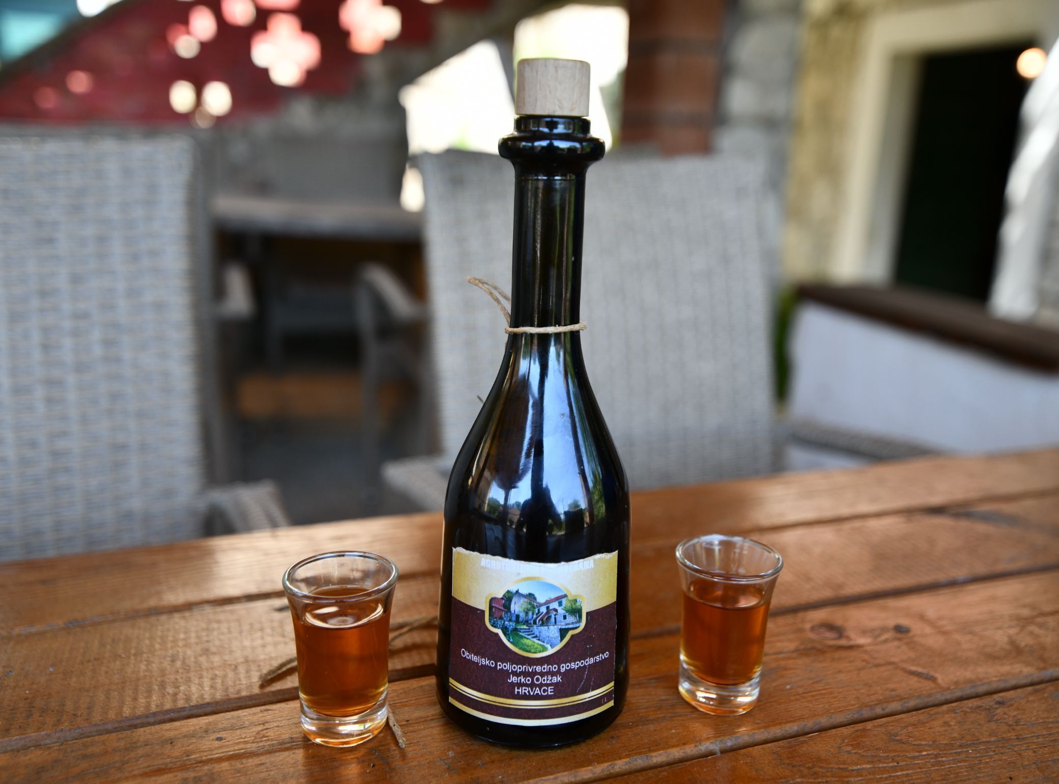 Une bouteille de produit de l’exploitation agricole familiale Obiteljski poljoprivredno gospodarstvo Jerko Odzak, originaire de Croatie, est posée sur une table en bois, accompagnée de deux petits verres à liqueur.