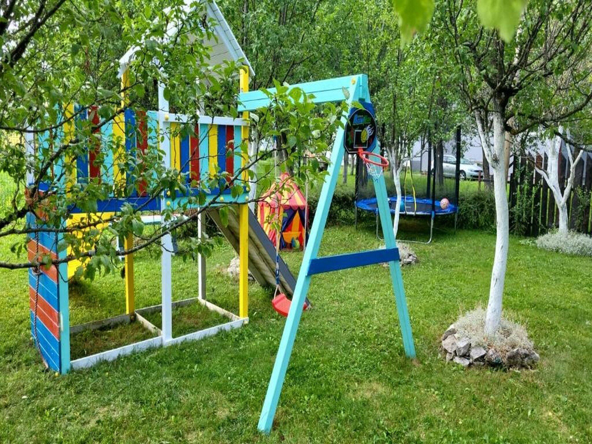Un parque infantil con una estructura de juegos de colores, que incluye un tobogán y columpios, rodeado de césped y árboles.