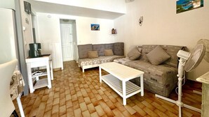 A compact living space with a sofa, a coffee table, a small table with a coffee maker, and a fan. There is a door leading to another room and a wall-mounted air conditioner. - Vacations in Corsica (Santa-Maria-Poggio)