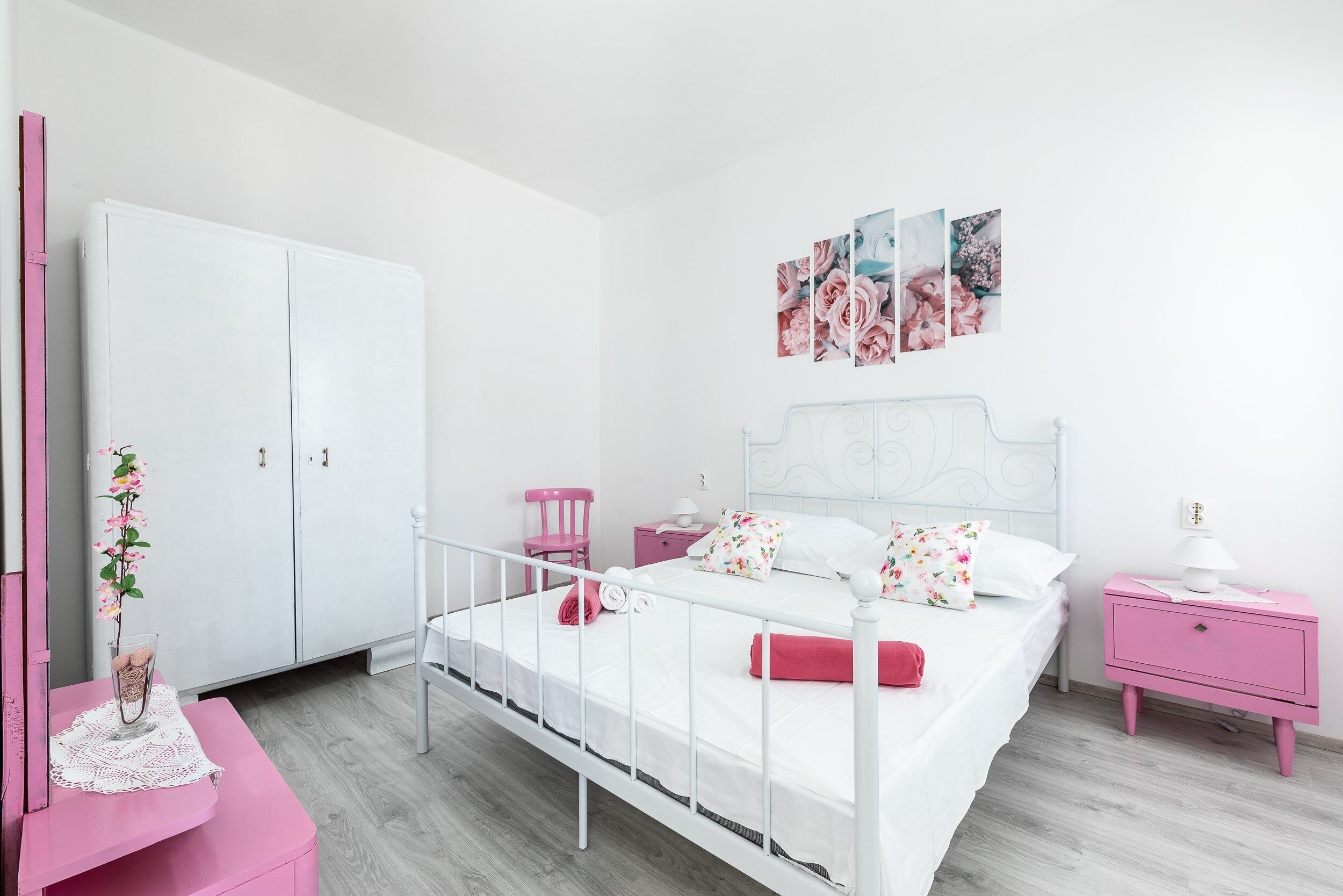 Un dormitorio con una cama blanca, mesitas de noche rosas, un armario blanco y decoración floral en la pared.