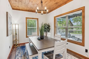 A dining area with a wooden table, white chairs, a chandelier, and large windows with blinds.