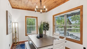 A dining area with a wooden table, white chairs, a chandelier, and large windows with blinds. - Luxury Cabin|Indoor Pool|Hot Tub|Ping Pong (Sevierville)