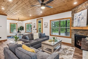 A spacious living room with a fireplace, large windows, and a ceiling fan.