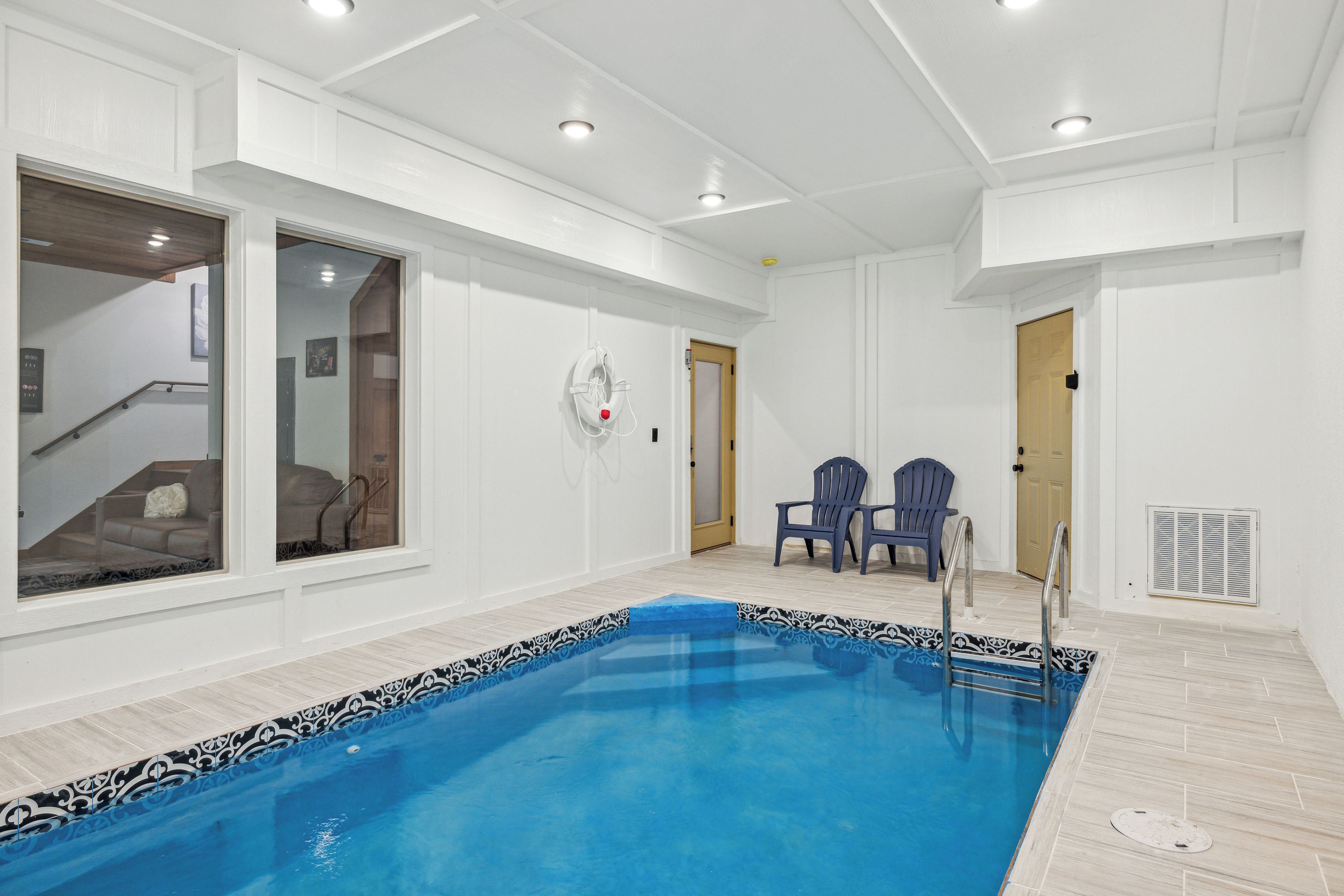A spacious indoor swimming pool with clear blue water, surrounded by white walls and a patterned pool edge. There are two chairs near the pool and a door leading to another room.