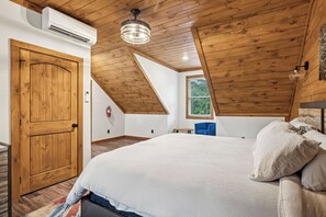 Um quarto com teto de madeira, uma cama com roupa de cama branca, uma cadeira azul e uma janela com vista para vegetação.