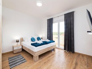A bedroom with a wooden bed, a nightstand, a black and white striped rug, and a view through a large window with dark curtains. - Apartments Mistral (Razanac)