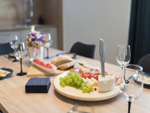 A dining table with a plate of cheese, grapes, and a knife. There are glasses of wine and a vase with flowers in the background. - Apartments Mistral (Razanac)