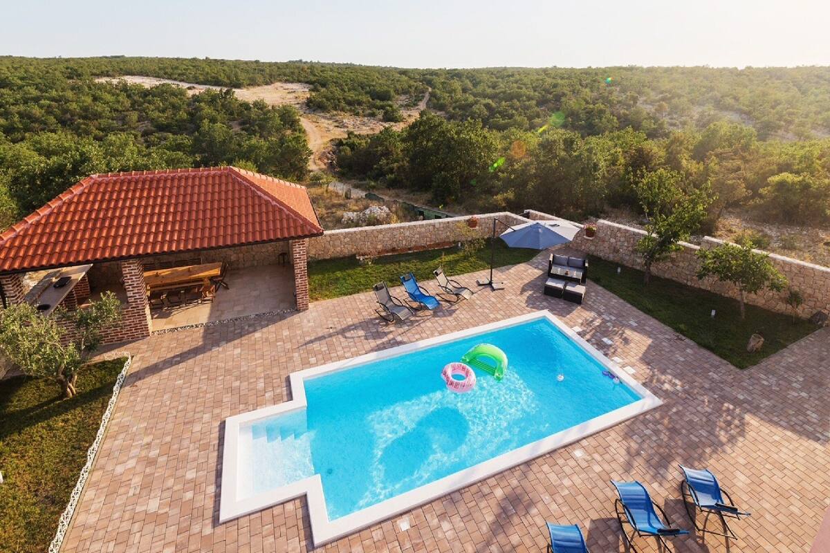 A swimming pool with lounge chairs and a barbecue area, surrounded by a stone wall and lush greenery.