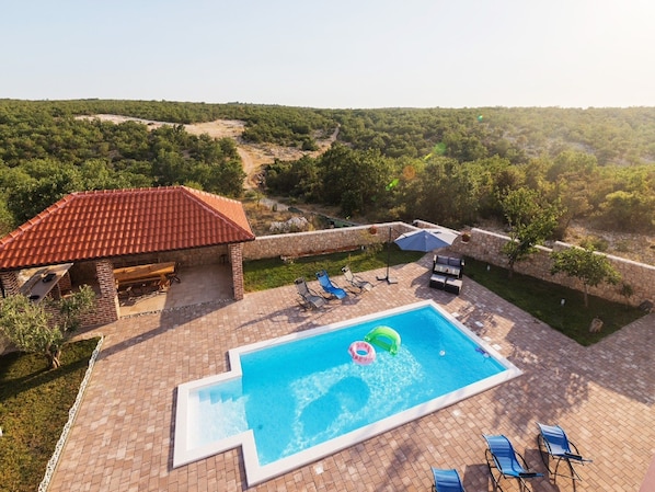 A swimming pool with lounge chairs and a barbecue area, surrounded by a stone wall and lush greenery. - Apartments Mistral (Razanac)